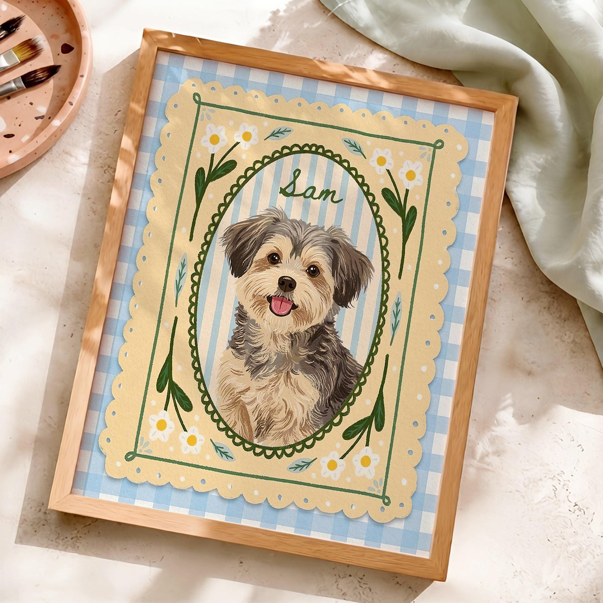 A framed illustration of a small, fluffy dog with brown and grey fur, tongue out, centered in an oval frame. The background features a light blue and white gingham pattern, with a yellow scalloped border decorated with white flowers.