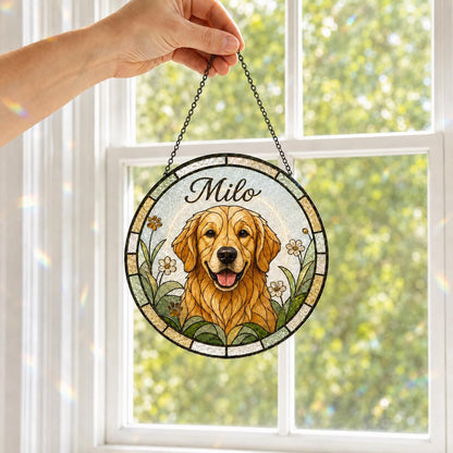 A circular stained glass suncatcher featuring a golden retriever illustration with the name Milo printed above. The artwork is surrounded by green foliage and small white flowers, framed by a dark border and suspended by a metal chain.