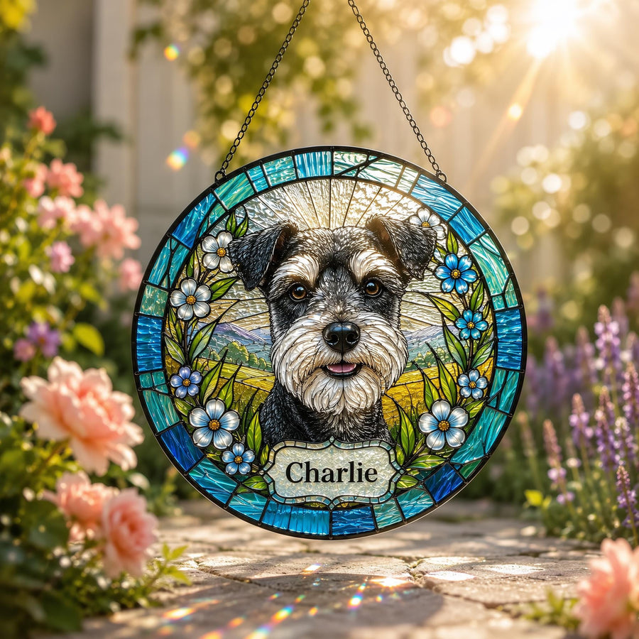 A circular stained glass suncatcher featuring a portrait of a black and gray Schnauzer dog. The dog is surrounded by blue flowers and green leaves. A white rectangular plaque at the bottom displays the name Charlie in black text.