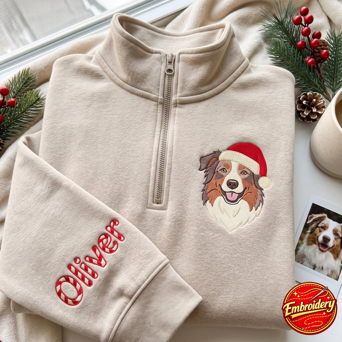 A beige quarter-zip sweatshirt featuring an embroidered Australian Shepherd wearing a red and white Santa hat on the chest. The sleeve is embroidered with the name Oliver in a red and white candy cane striped font style.