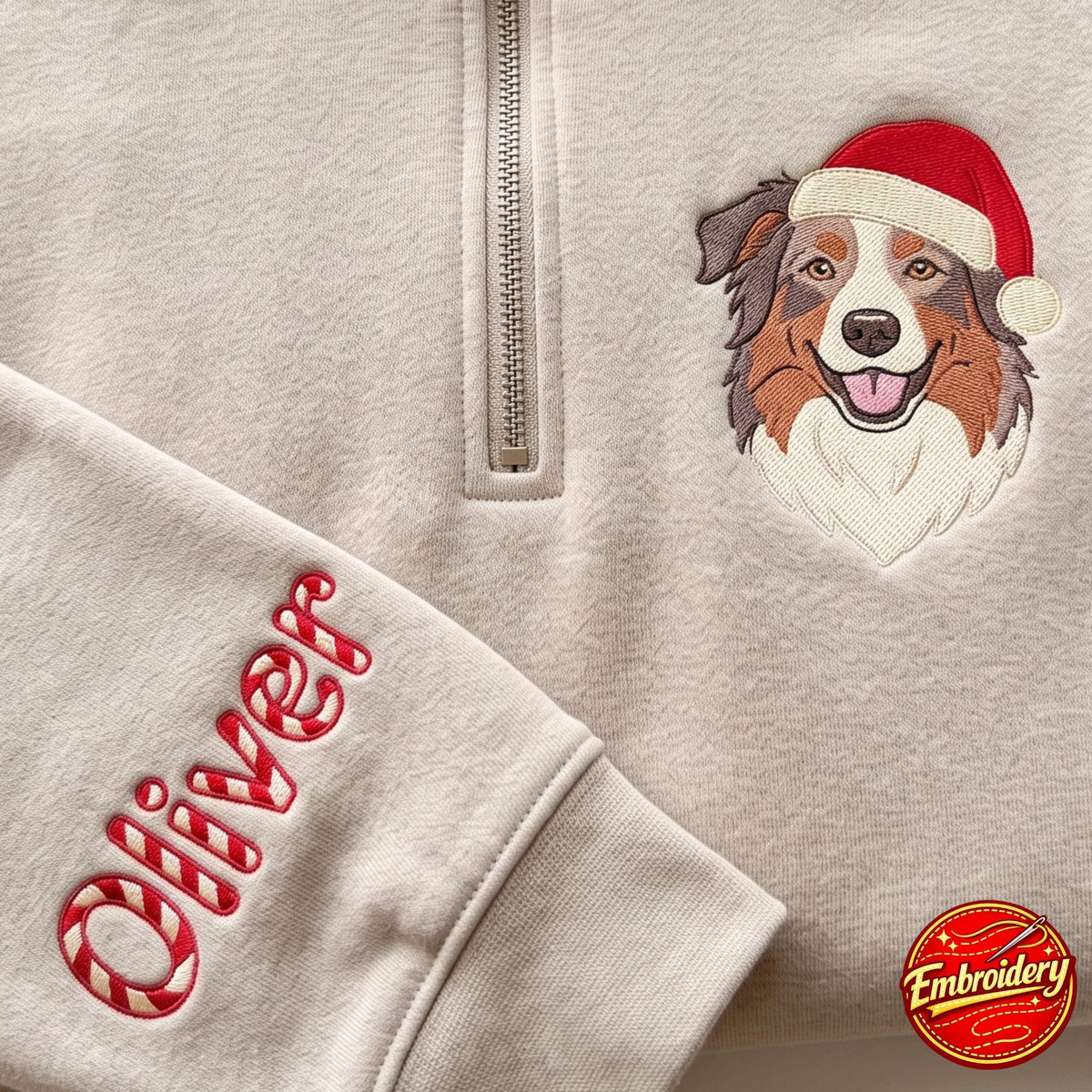 A beige quarter-zip sweatshirt featuring an embroidered Australian Shepherd wearing a red Santa hat. The sleeve is personalized with the name Oliver in a red and white candy cane striped font. A small circular embroidery logo is visible.
