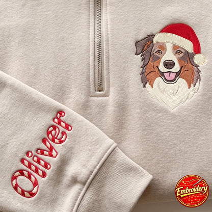 A beige quarter-zip sweatshirt featuring an embroidered Australian Shepherd wearing a red Santa hat. The sleeve is personalized with the name Oliver in a red and white candy cane striped font. A small circular embroidery logo is visible.