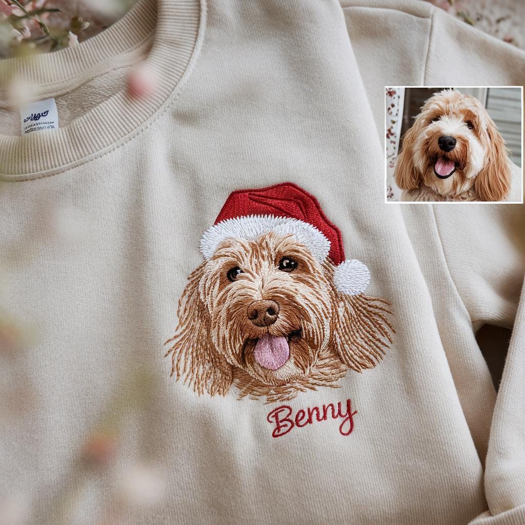 Cream-colored sweatshirt featuring an embroidered image of a dog wearing a red Santa hat. The dog's name, "Benny," is embroidered in red script below the image. A smaller image of the dog is in the upper right corner.