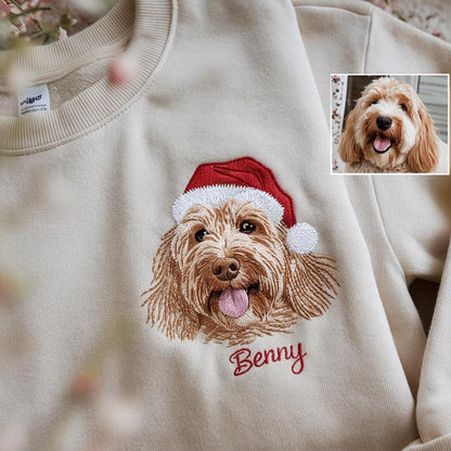Cream-colored sweatshirt featuring an embroidered image of a dog wearing a red Santa hat. The dog's name, "Benny," is embroidered in red script below the image. A smaller image of the dog is in the upper right corner.