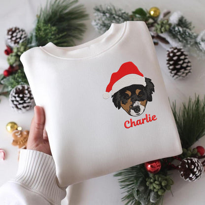 White sweatshirt featuring an embroidered dog wearing a Santa hat. The dog's face is black, brown, and tan, with the name "Charlie" embroidered in red below. Festive holiday apparel.