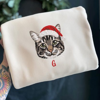 Cream-colored sweatshirt featuring an embroidered cat face wearing a red and white Santa hat. The cat has gray and brown tabby markings and green eyes. A red letter "G" is embroidered below the cat's face.