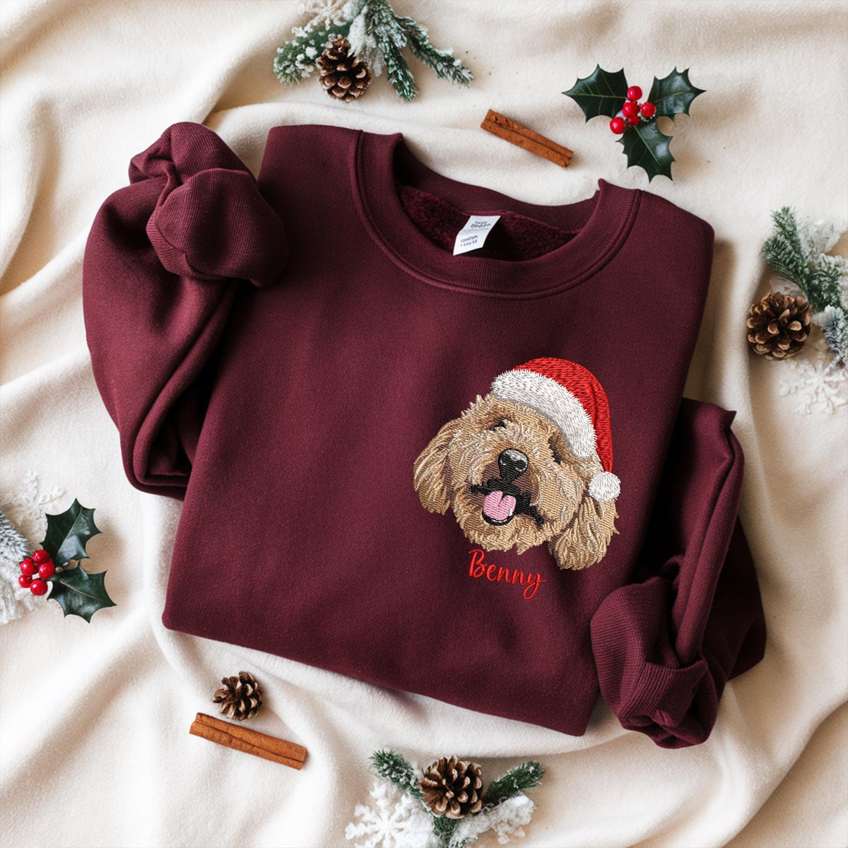Burgundy sweatshirt with an embroidered dog wearing a Santa hat. The dog's name, "Benny," is embroidered in red below the dog. The sweatshirt is displayed on a cream-colored surface with holiday decorations.