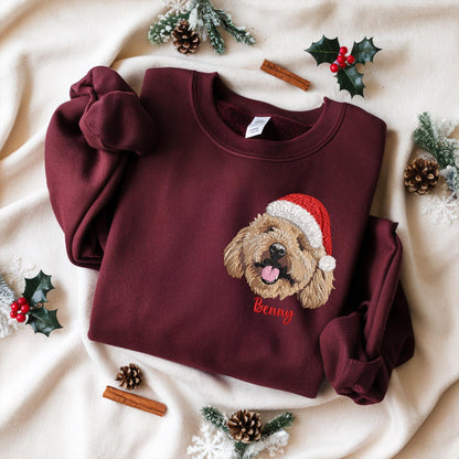 Burgundy sweatshirt with an embroidered dog wearing a Santa hat. The dog's name, "Benny," is embroidered in red below the dog. The sweatshirt is displayed on a cream-colored surface with holiday decorations.