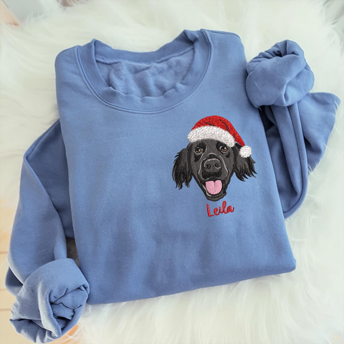 A light blue sweatshirt featuring an embroidered black dog wearing a red and white Santa hat. The dog's name, "Leila," is embroidered in red script below the dog's face. The sweatshirt is folded on a white, fluffy surface.