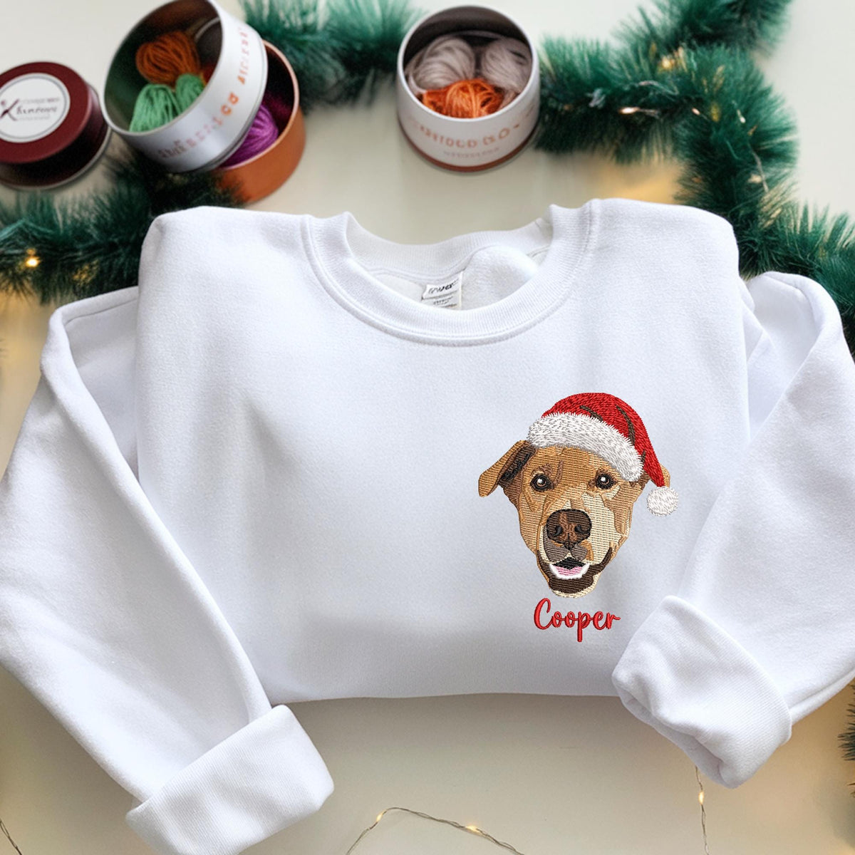 White sweatshirt featuring an embroidered dog wearing a Santa hat. The dog's name, "Cooper," is embroidered in red below the image. The sweatshirt has long sleeves and a crew neck, perfect for the holiday season.