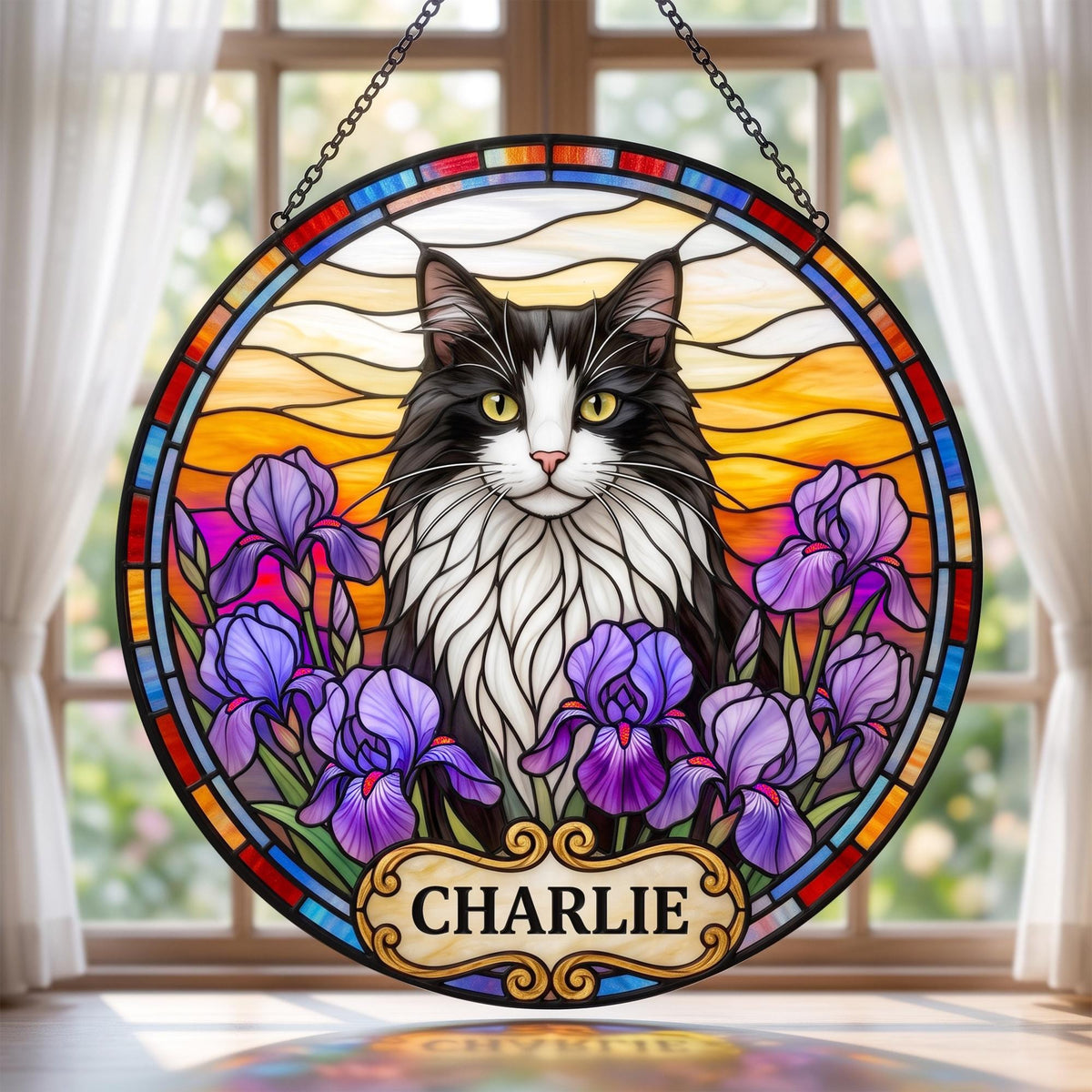 A circular stained glass suncatcher featuring a black and white cat with yellow eyes surrounded by purple iris flowers. The background shows a sunset sky with orange and yellow hues. A decorative plaque at the bottom reads CHARLIE.