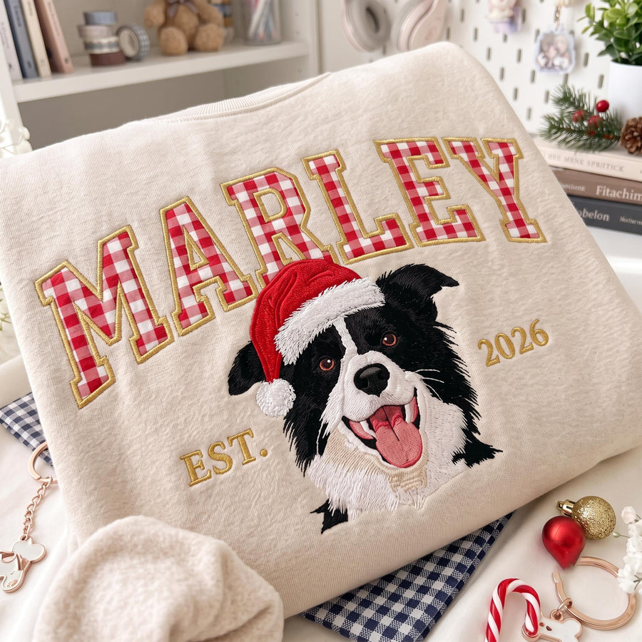 A beige sweatshirt featuring the embroidered name MARLEY in red and white gingham letters. Below the name is an embroidered black and white dog wearing a red Santa hat, with the text EST. and 2026 in gold thread on either side.