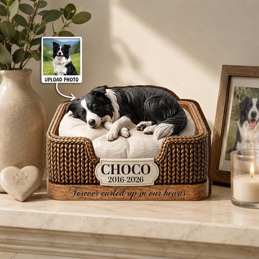 A memorial figurine of a black and white dog sleeping on a cream-colored cushion inside a brown woven-textured bed. A plaque on the front reads CHOCO 2016-2026, with the text Forever curled up in our hearts engraved below it.