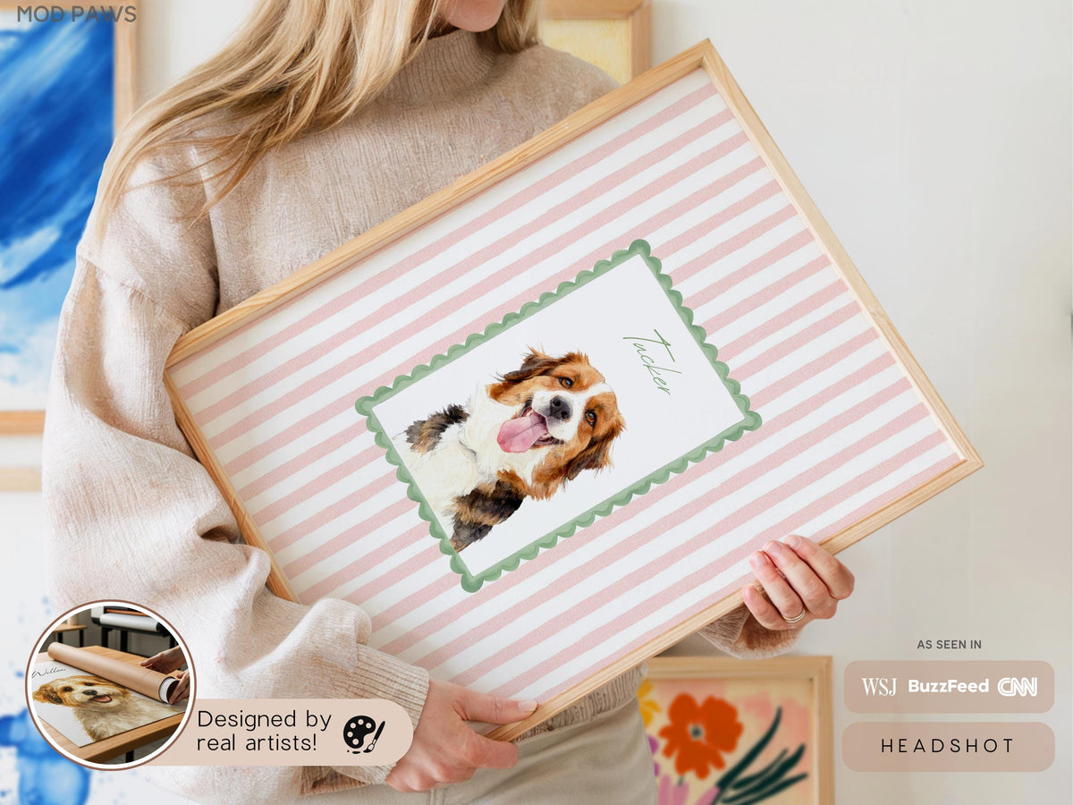 A framed print featuring a watercolor illustration of a dog named Tucker, set against a pink and white striped background. The print is in a light wood frame. The text "Designed by real artists!" is visible.