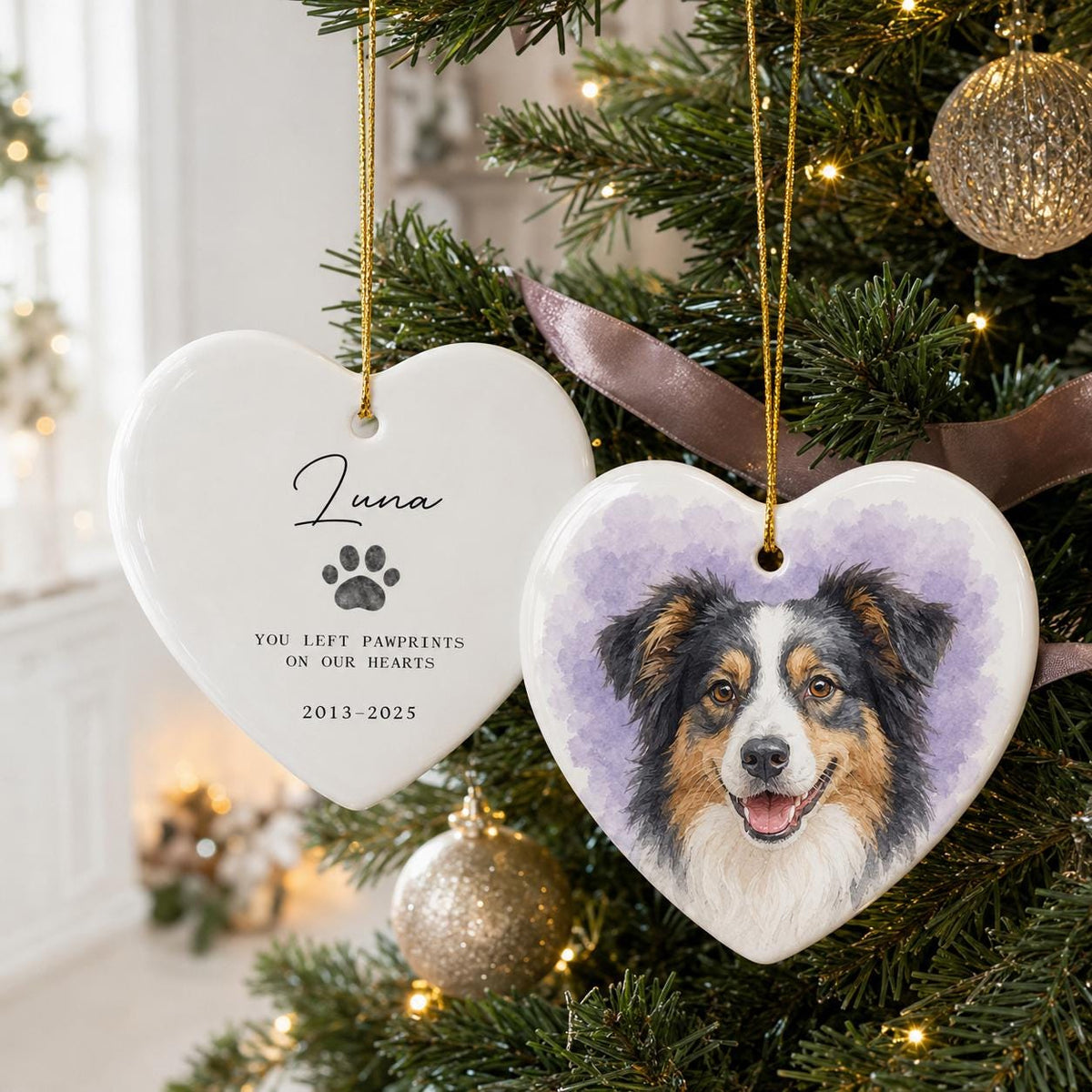 Two white heart-shaped ceramic ornaments hanging by gold strings. One features a watercolor portrait of a tricolor dog against a purple background. The other displays the name Luna, a paw print, and the text: YOU LEFT PAWPRINTS ON OUR HEARTS 2013-2025.