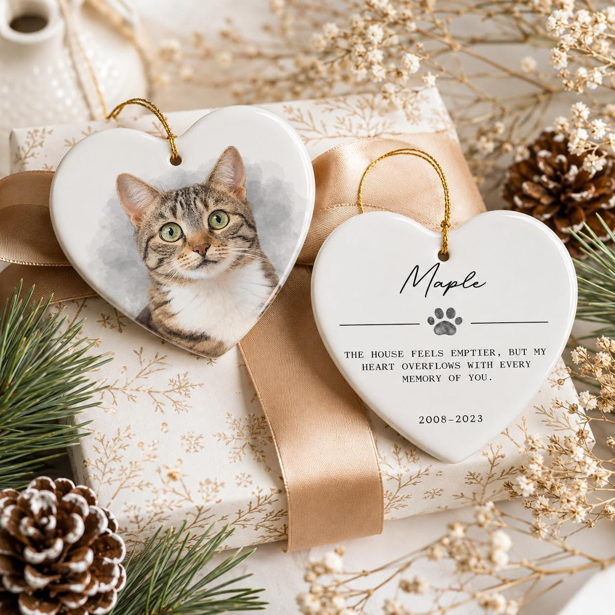 Two white heart-shaped ceramic ornaments with gold hanging strings. One ornament features a portrait of a brown tabby cat. The other ornament displays the name Maple, a paw print icon, and the text: The house feels emptier, but my heart overflows with every memory of you. 2008-2023.