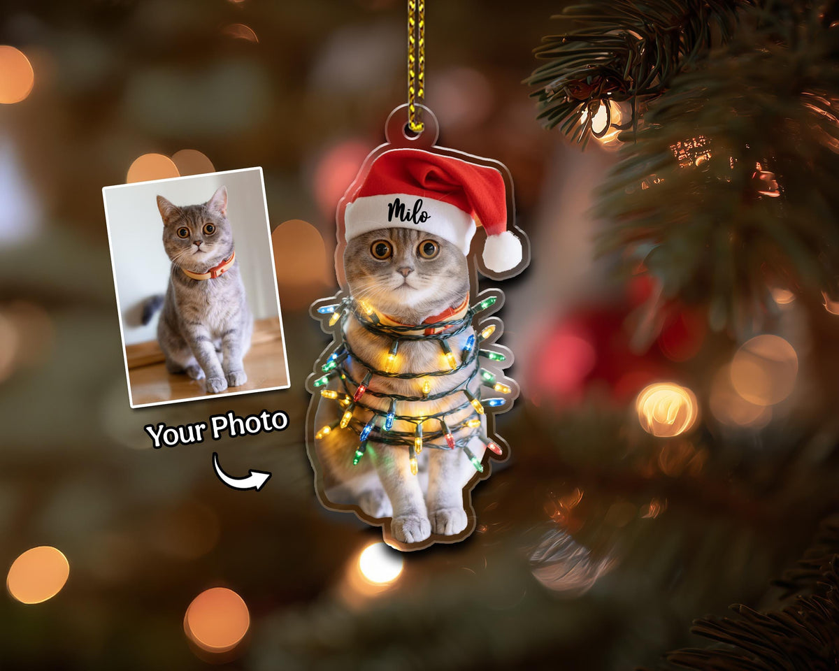 A clear acrylic Christmas ornament featuring a cat wearing a Santa hat with the name "Milo". The cat is wrapped in string lights. A photo of the cat is on a separate card. The words "Your Photo" are printed on the card.