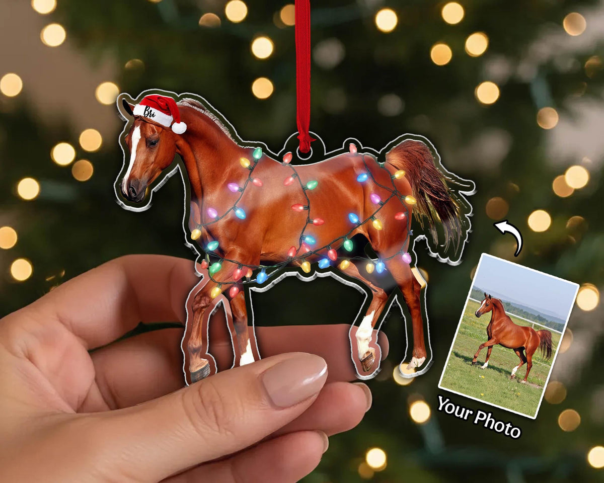 A brown horse ornament with a Santa hat and Christmas lights. The horse is wearing a red Santa hat and is decorated with colorful lights. A small photo of a horse is in the lower right corner. The ornament is hanging from a red ribbon.