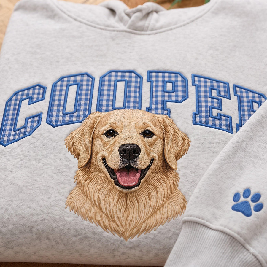 A light gray hooded sweatshirt featuring an embroidered golden retriever portrait centered on the chest. Above the dog, the name COOPER is stitched in blue and white gingham block letters. A blue paw print is embroidered on the sleeve.