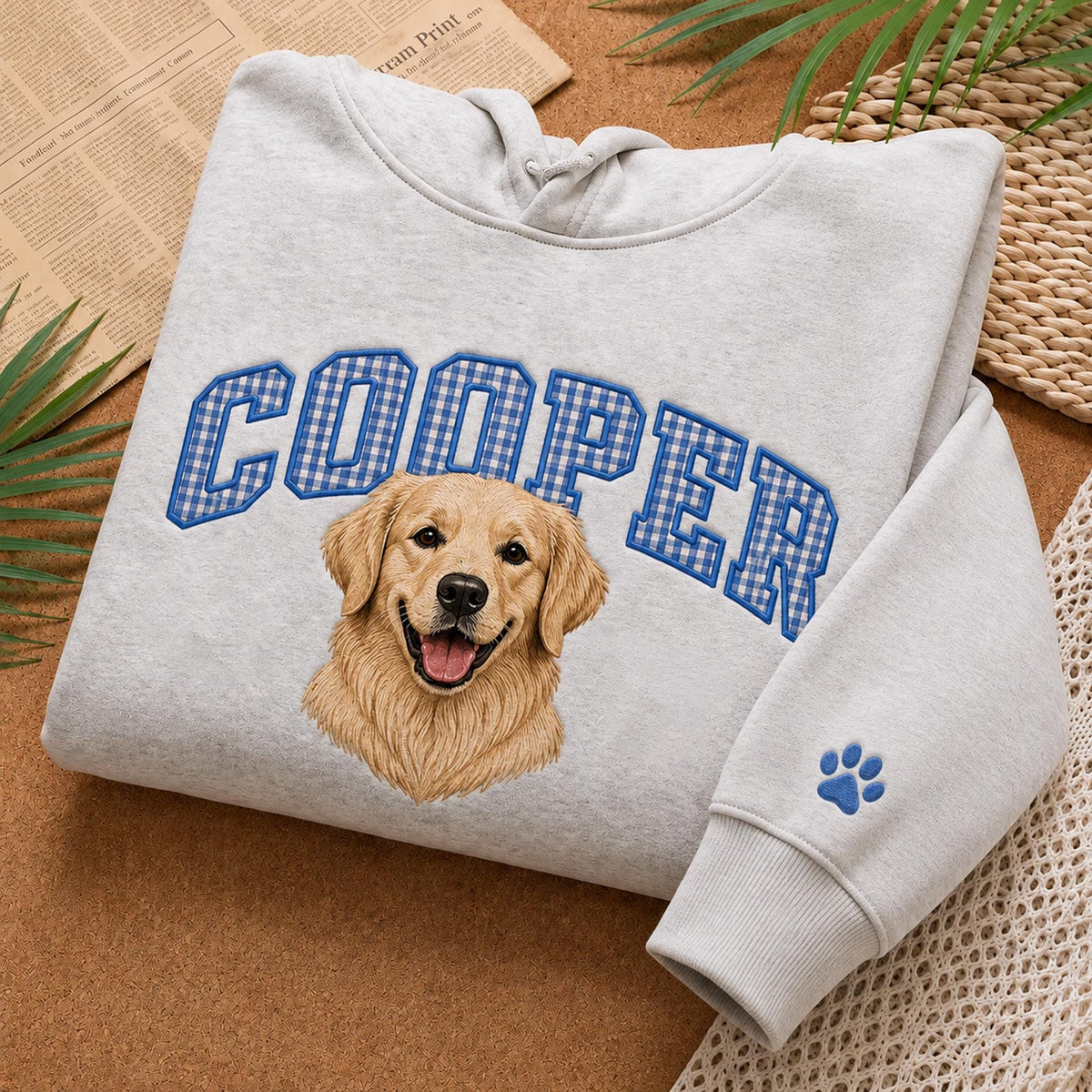 A light gray hooded sweatshirt featuring the name COOPER in blue and white gingham block letters arched over a realistic illustration of a golden retriever dog. A small blue paw print is embroidered on the sleeve cuff.