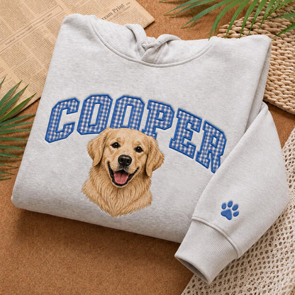 A light gray hooded sweatshirt featuring the name COOPER in blue and white gingham block letters arched over a realistic illustration of a golden retriever dog. A small blue paw print is embroidered on the sleeve cuff.