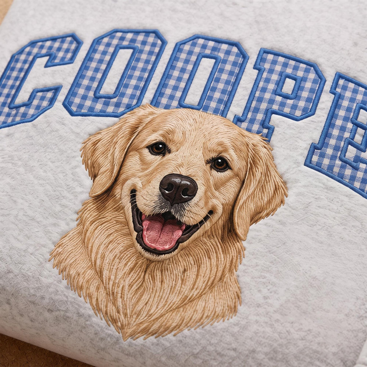 A white textured fabric featuring an embroidered golden retriever portrait with a smiling expression and open mouth. Above the dog, the name COOPER is stitched in large block letters with a blue and white gingham checkered pattern.