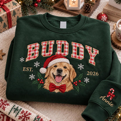 A dark green crewneck sweatshirt featuring an embroidered golden retriever wearing a Santa hat and a red bow tie. The text BUDDY is in red and white gingham letters above the dog, with EST. 2026 and white snowflakes. The sleeve says Annie.