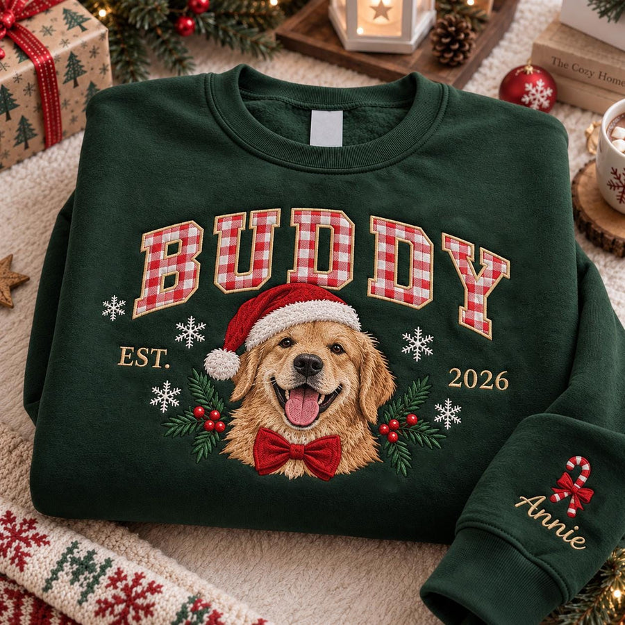 A dark green crewneck sweatshirt featuring an embroidered golden retriever wearing a Santa hat and a red bow tie. The text BUDDY is in red and white gingham letters above the dog, with EST. 2026 and white snowflakes. The sleeve says Annie.