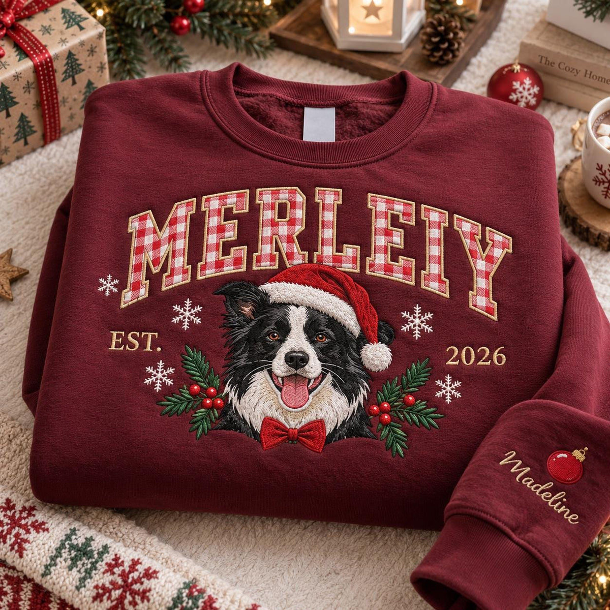 A maroon crewneck sweatshirt featuring the embroidered text MERLEIY in a red and white gingham pattern. Below, a border collie wearing a Santa hat and red bow tie is surrounded by snowflakes, holly, and the text EST. 2026. The sleeve features the name Madeline and a red ornament.