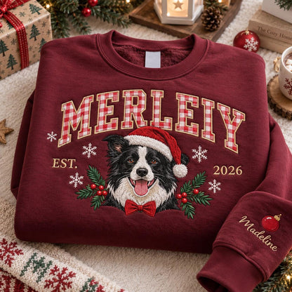 A maroon crewneck sweatshirt featuring the embroidered text MERLEIY in a red and white gingham pattern. Below, a border collie wearing a Santa hat and red bow tie is surrounded by snowflakes, holly, and the text EST. 2026. The sleeve features the name Madeline and a red ornament.