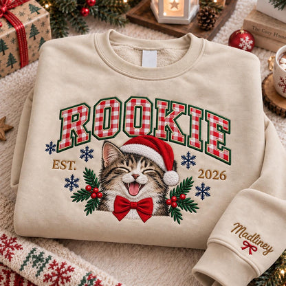A beige crewneck sweatshirt featuring the word ROOKIE in red and white gingham letters. Below, an embroidered tabby cat wears a red Santa hat and a red bow tie, flanked by holly and snowflakes. The text EST. 2026 appears, with Madliney on the sleeve.