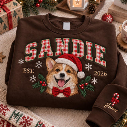 A dark brown crewneck sweatshirt featuring the name SANDIE in red and white gingham block letters. Below, an embroidered Corgi wears a Santa hat and red bow tie, flanked by holly and snowflakes. EST. 2026 is printed, with Jame and a candy cane on the sleeve.