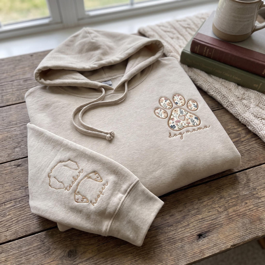 A beige hooded sweatshirt featuring an embroidered floral paw print design on the chest with the text "dog mama" below it. The sleeve is embroidered with two dog head outlines and the names "tucker" and "hooper" in cursive script.