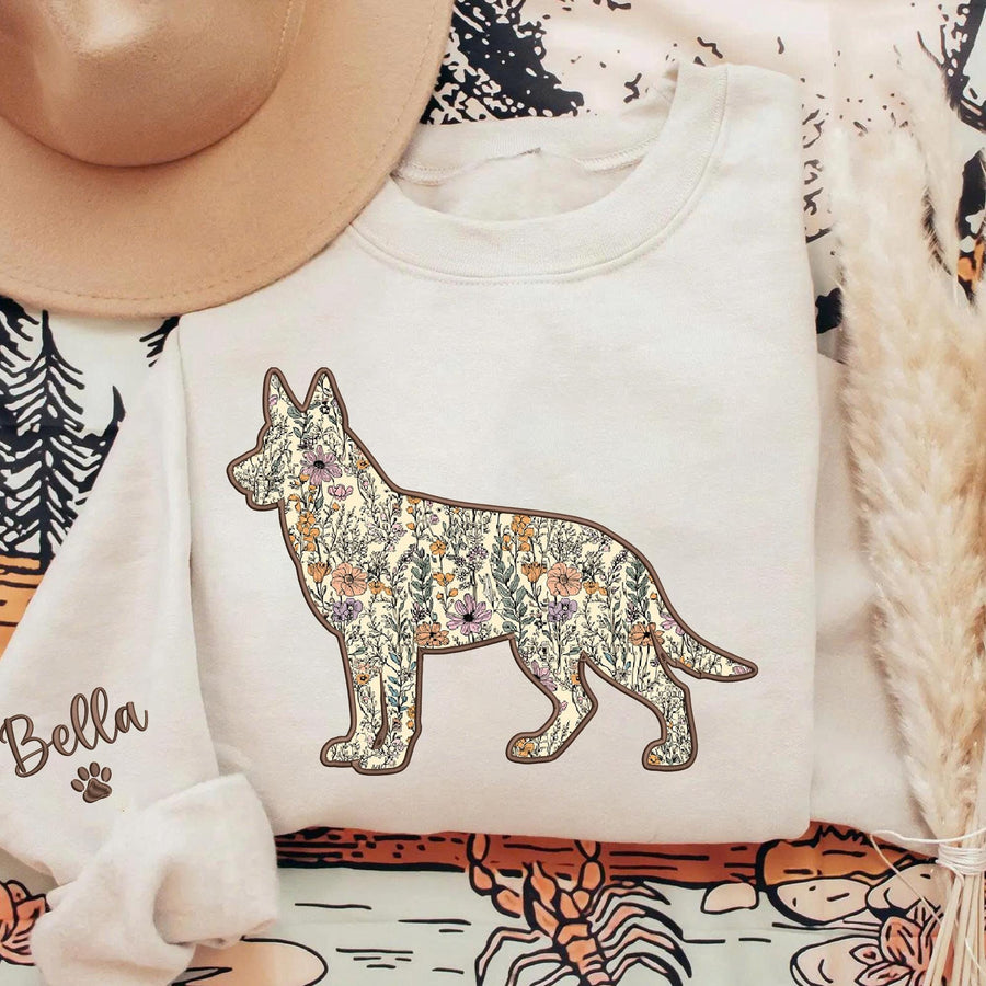 A cream-colored crewneck sweatshirt featuring a silhouette of a German Shepherd dog filled with a colorful floral pattern. The sleeve displays the name Bella in cursive script next to a small paw print icon.
