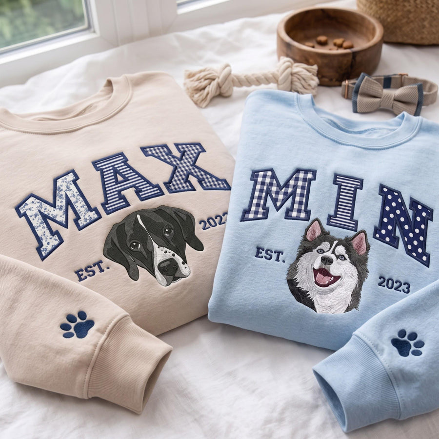 A beige sweatshirt with the embroidered name MAX and a black and white dog portrait, and a light blue sweatshirt with the embroidered name MIN and a husky portrait. Both feature paw print sleeve details and the text EST. 2023.