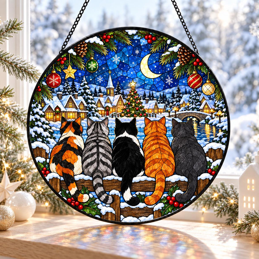 A circular stained glass suncatcher featuring five cats sitting on a wooden fence, overlooking a snowy village at night. The scene includes a crescent moon, a decorated Christmas tree, pine branches, and hanging ornaments in a vibrant, multi-colored mosaic style.
