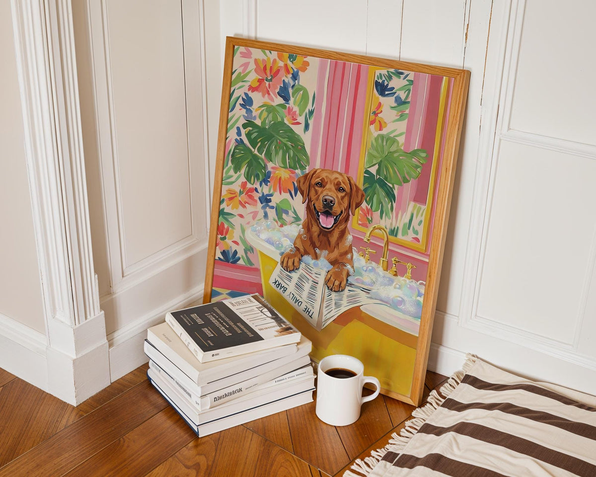 A framed art print featuring a brown Labrador Retriever in a yellow bathtub, reading "The Daily Bark" newspaper. The artwork has a floral and striped background. A stack of books and a coffee mug are in the foreground.