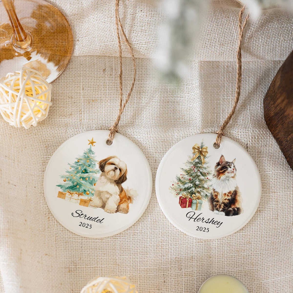 Two white ceramic Christmas ornaments with pet portraits. One features a Shih Tzu dog and the name "Strudel" with the year 2025. The other shows a cat and the name "Hershey" with the year 2025. Both have a Christmas tree design.
