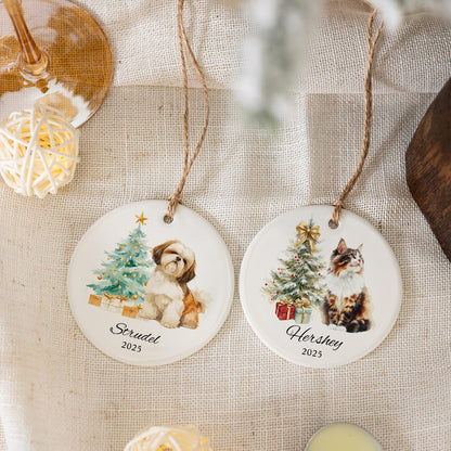 Two white ceramic Christmas ornaments with pet portraits. One features a Shih Tzu dog and the name "Strudel" with the year 2025. The other shows a cat and the name "Hershey" with the year 2025. Both have a Christmas tree design.