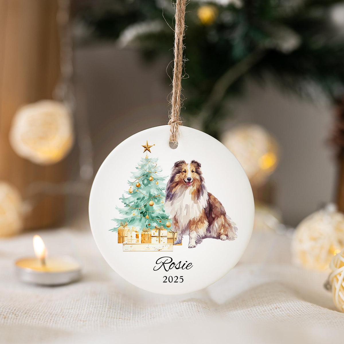 A round, white Christmas ornament with a watercolor illustration of a Sheltie dog next to a decorated Christmas tree and wrapped gifts. The ornament has the name "Rosie" and the year "2025" printed on it, and is suspended by a twine string.