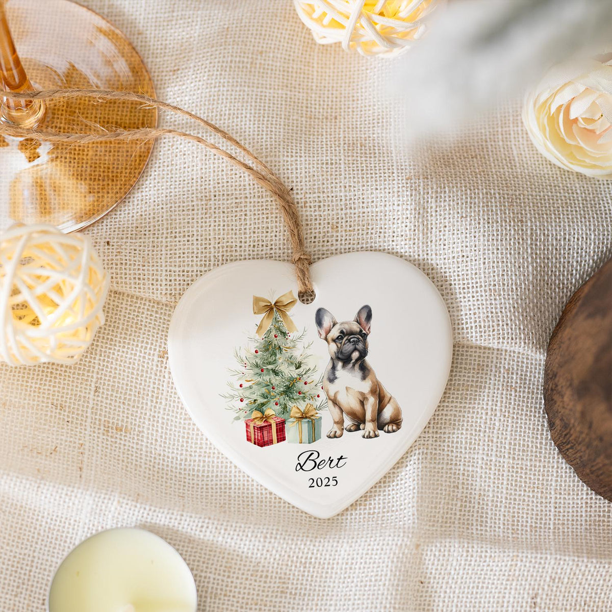 Heart-shaped ceramic Christmas ornament featuring a French Bulldog, Christmas tree, and wrapped gifts. The ornament has a jute string for hanging and includes the name "Bert" and the year "2025".
