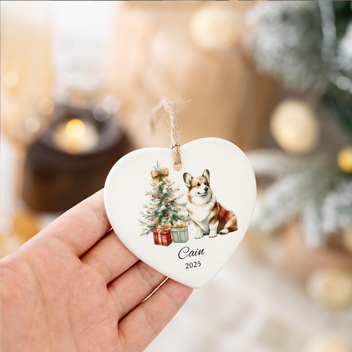 Heart-shaped ceramic Christmas ornament featuring a watercolor illustration of a Corgi dog, Christmas tree, and gifts. The ornament has a twine loop for hanging and includes the name "Cain" and the year "2025".