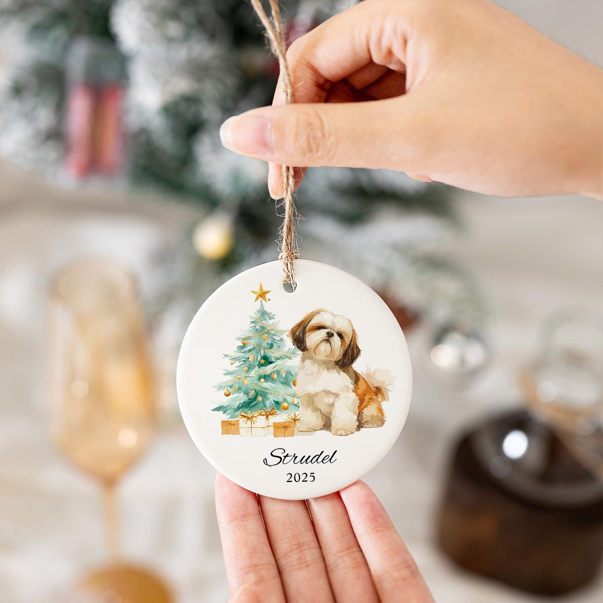 A round, white Christmas ornament featuring a watercolor illustration of a Shih Tzu dog, a decorated Christmas tree, and wrapped gifts. The ornament has the name "Strudel" and the year "2025" printed on it.