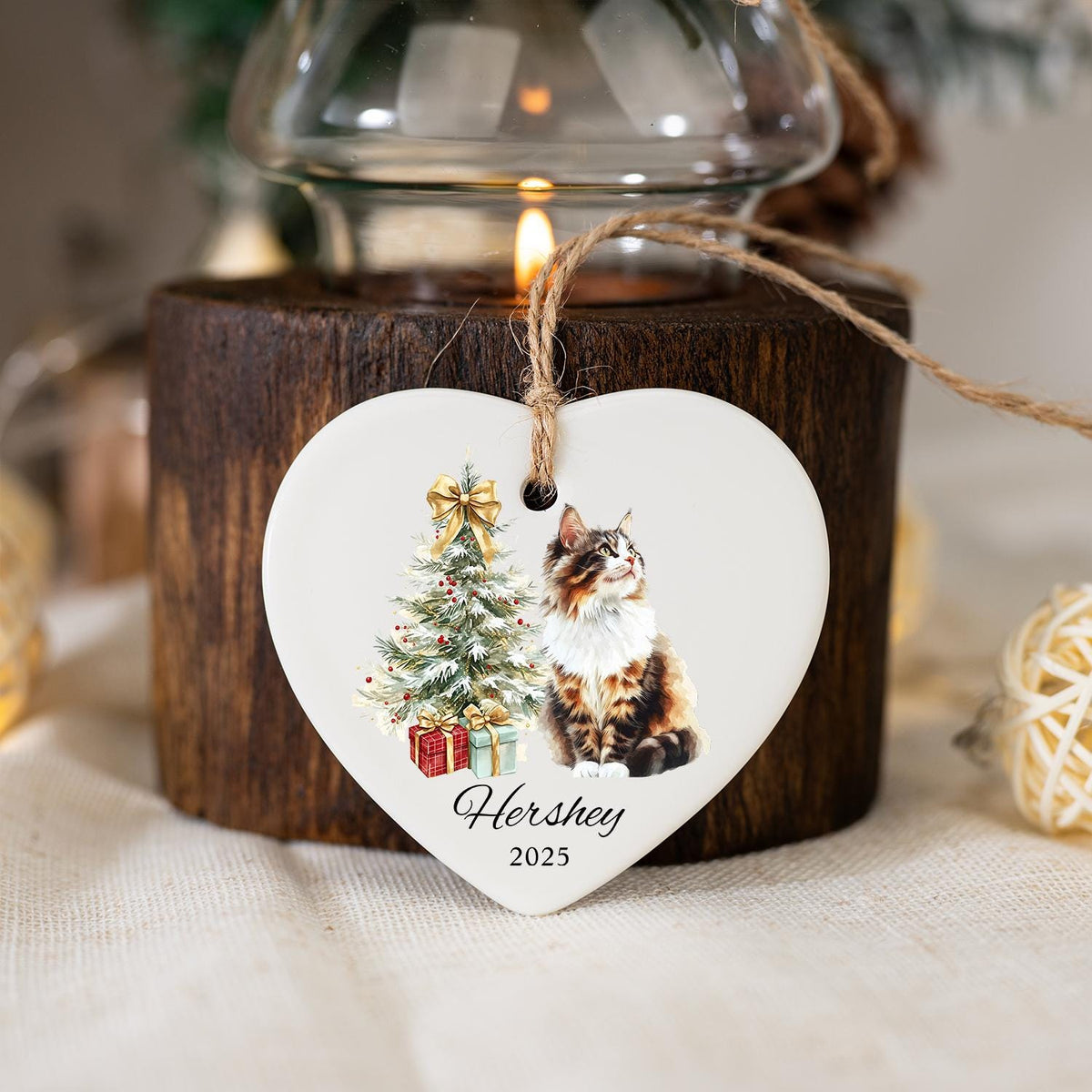 Heart-shaped ceramic Christmas ornament featuring a cat and a decorated Christmas tree with gifts. The ornament has the name "Hershey" and the year "2025" printed on it. It hangs from a natural twine string.