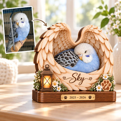 A wooden memorial plaque featuring carved angel wings cradling a blue and white budgie. The base includes a small lantern, white flowers, a paw print, and a gold plate engraved with the name Sky and the years 2025 - 2026.