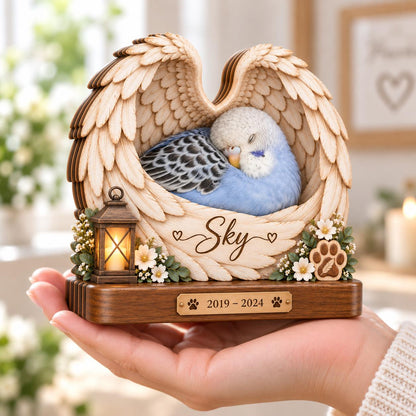 A wooden memorial plaque featuring a carved pair of angel wings cradling a sleeping blue and white budgerigar. The piece includes a small lantern, white flowers, a paw print, and the name Sky with the dates 2019-2024 engraved.