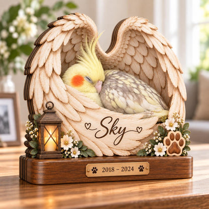 A wooden memorial sculpture featuring a sleeping cockatiel nestled within carved angel wings. The piece includes the name Sky, small white flowers, a miniature lantern, and a brass plaque engraved with the dates 2018 - 2024.