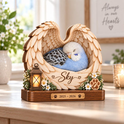 A wooden memorial sculpture featuring a sleeping blue and white budgie nestled within carved angel wings. The piece includes a small lantern, white flowers, a paw print, and the name Sky. A brass plaque displays the years 2025 - 2026.