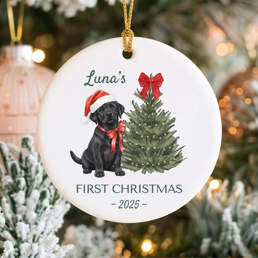 White ceramic Christmas ornament featuring a black Labrador puppy wearing a Santa hat and red bow. A green Christmas tree and the text "Luna's First Christmas - 2025" are also displayed.