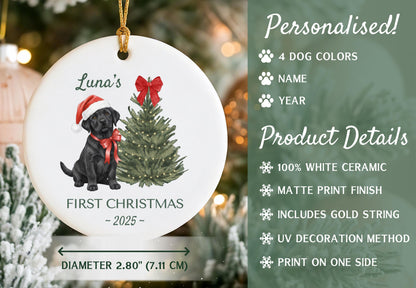 A white ceramic Christmas ornament with a black Labrador wearing a Santa hat and red bow, next to a Christmas tree. The ornament is 2.80 inches (7.11 cm) in diameter and has the text "Luna's First Christmas - 2025".